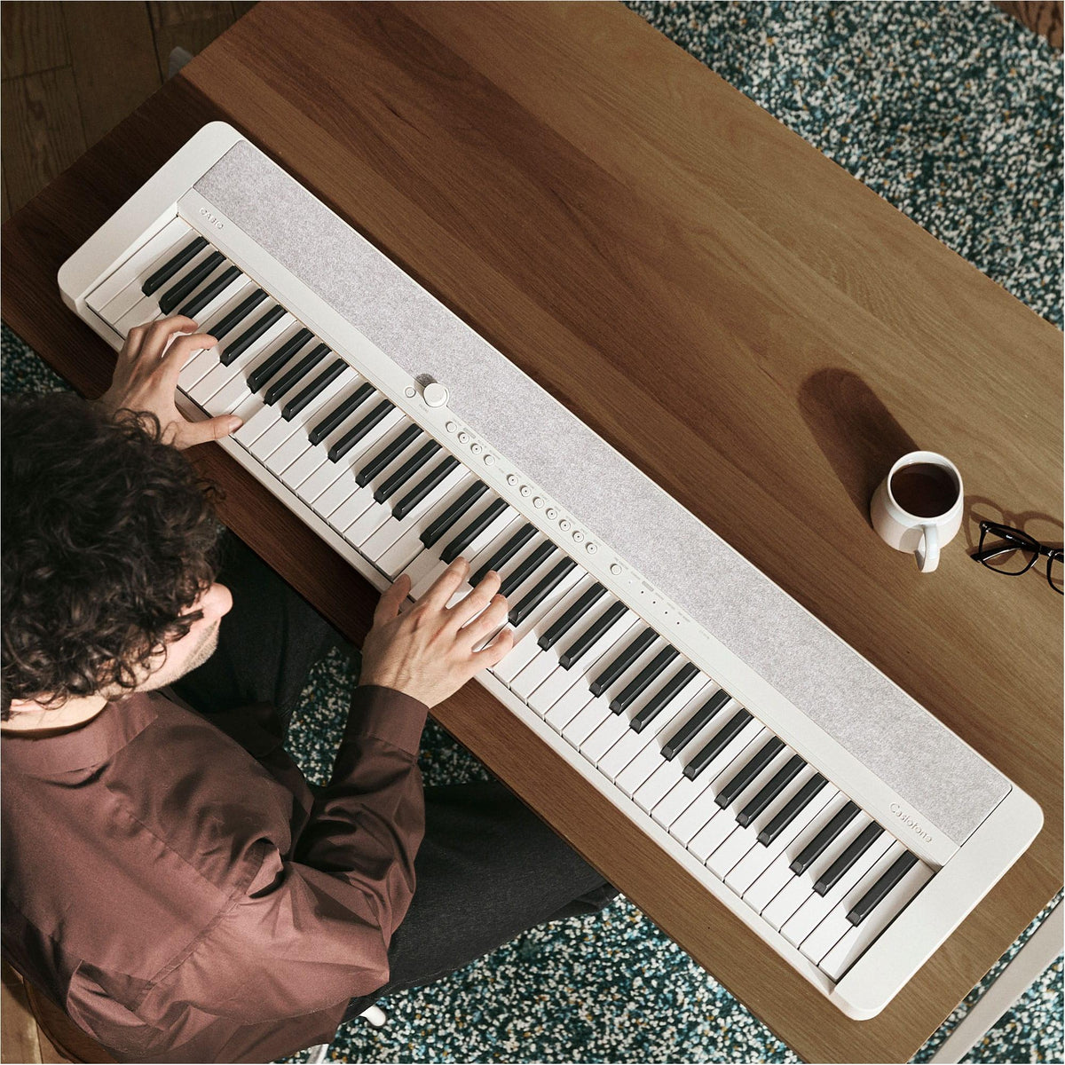 Overhead view of a person playing a white Casio CT-S1-76WE keyboard on a wooden desk beside a cup of coffee