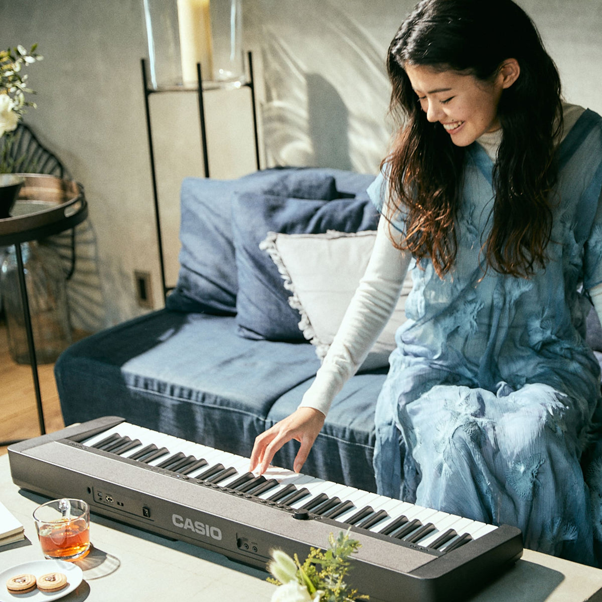 Casio CT-S1 keyboard on a coffee table in a living room, shown from the rear with the CASIO logo and connection jacks visible while a seated player smiles and plays the keys
